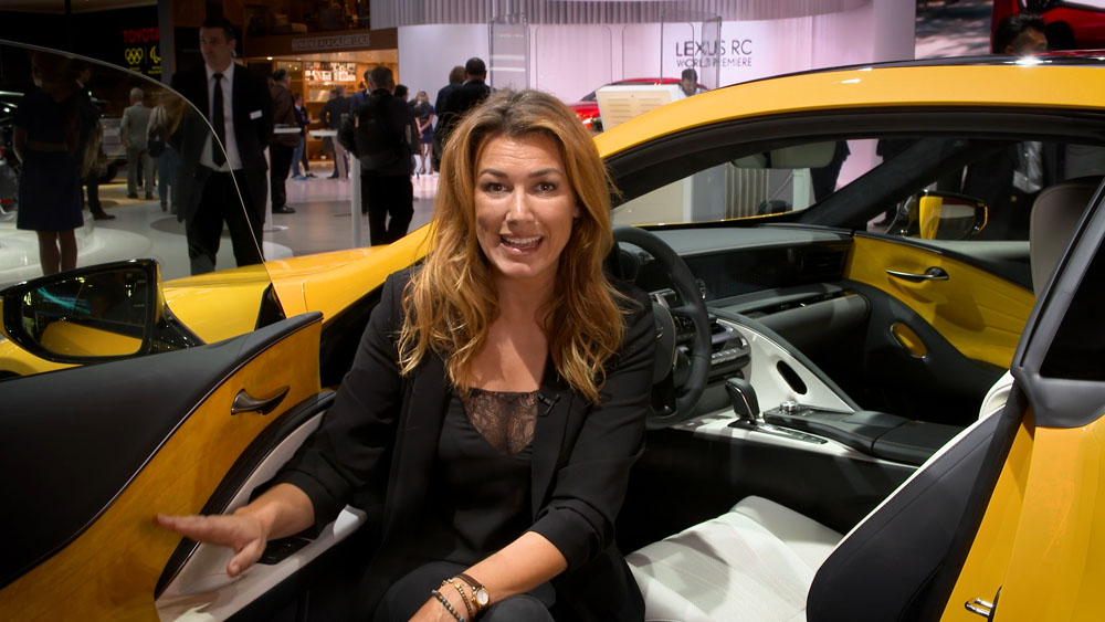 Lexus Europe Paris Reportage, video garage image, presenter Ilse De Vis in a low posture in front of the interior of a vibrant flare yellow paint finish Lexus LC 500h at the Paris Motor Show, members of the public and Michelin-starred chef, Andreas Møller in the background near a red Lexus RC